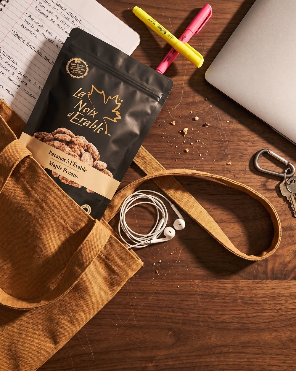 Un sac de pacanes à l'érable qui dépasse d'un tote, sur un bureau avec des notes, des surligneurs, un portable et des écouteurs